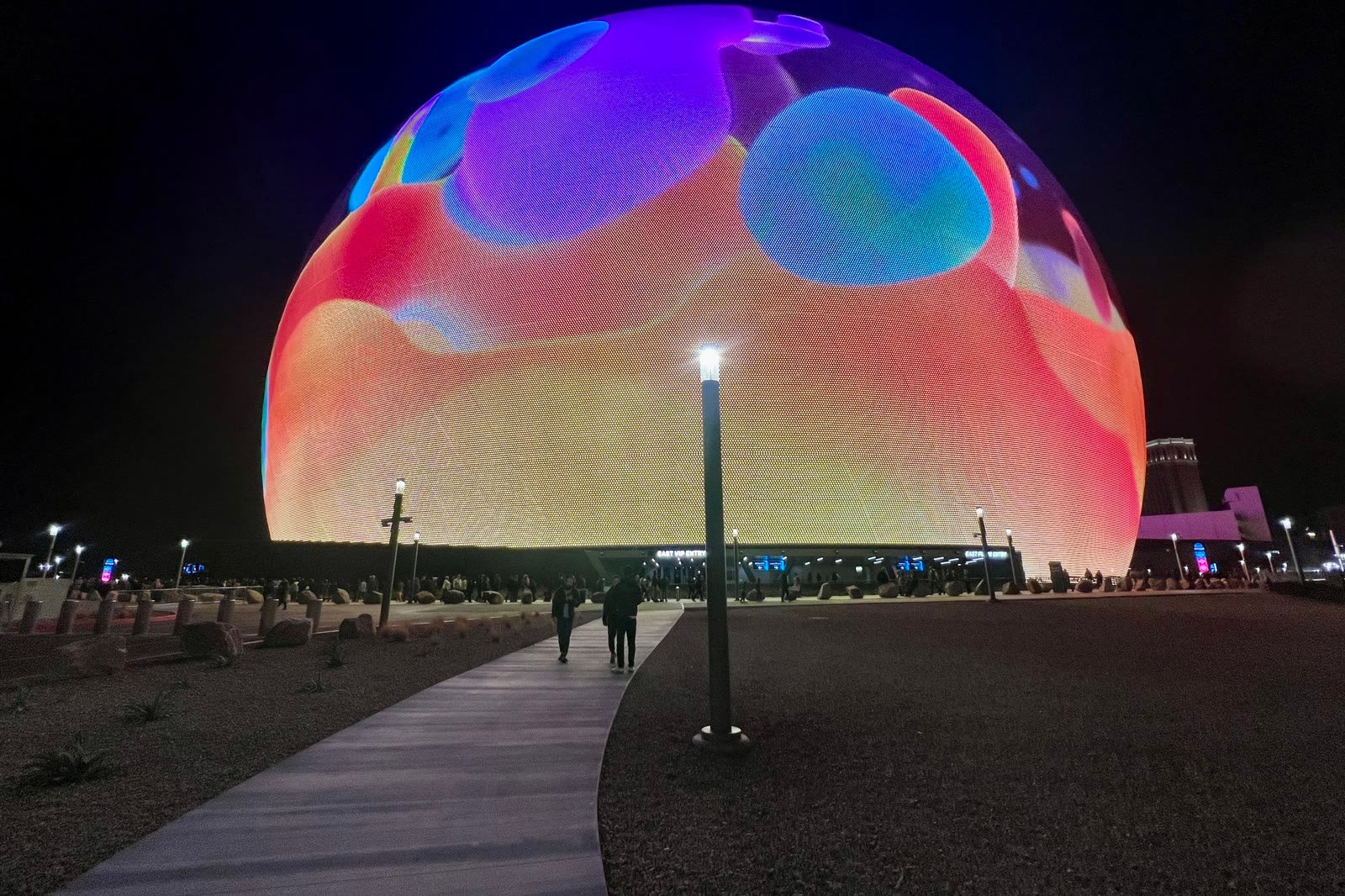 Sphere encendida de noche con manchas de colores, y gente que camina hacia ella por un sendero