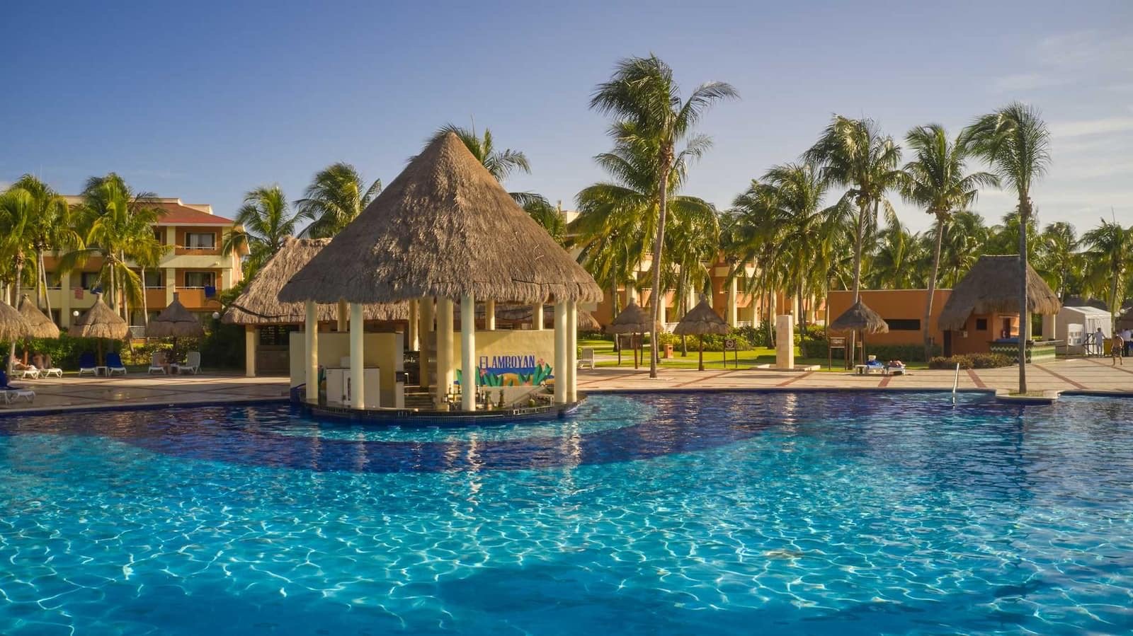 Alberca azul con un bar de palapa al centro y palmeras alrededor