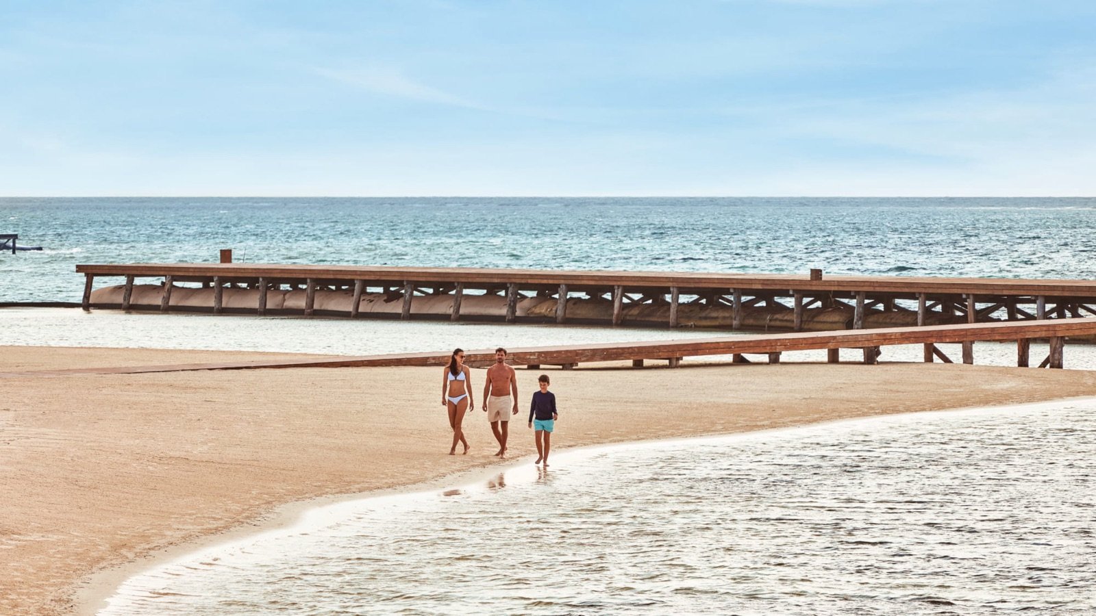Una familia camina por la orilla de una playa de arena blanca con un muelle al fondo
