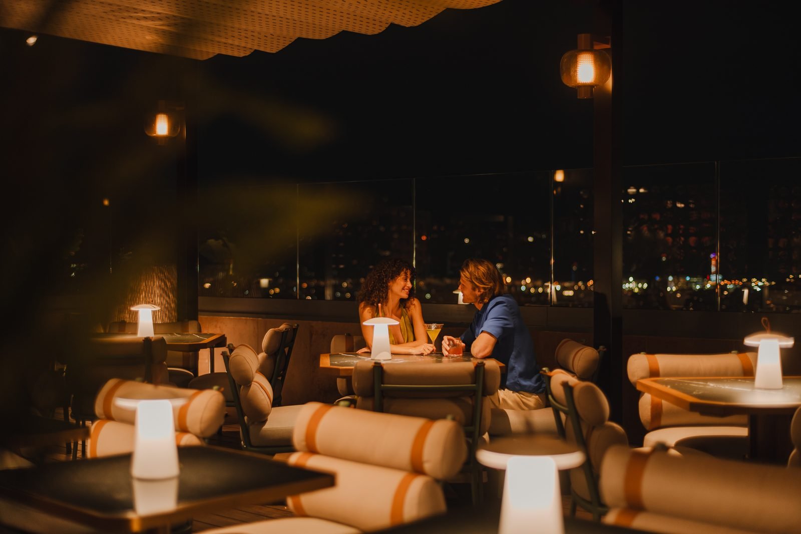 Una pareja brinda de noche en una terraza con lámparas de mesa y la ciudad iluminada
