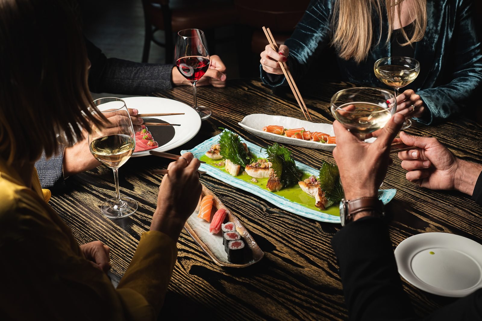 Varias manos con palillos alrededor de una mesa con sushi, sashimi y copas de vino