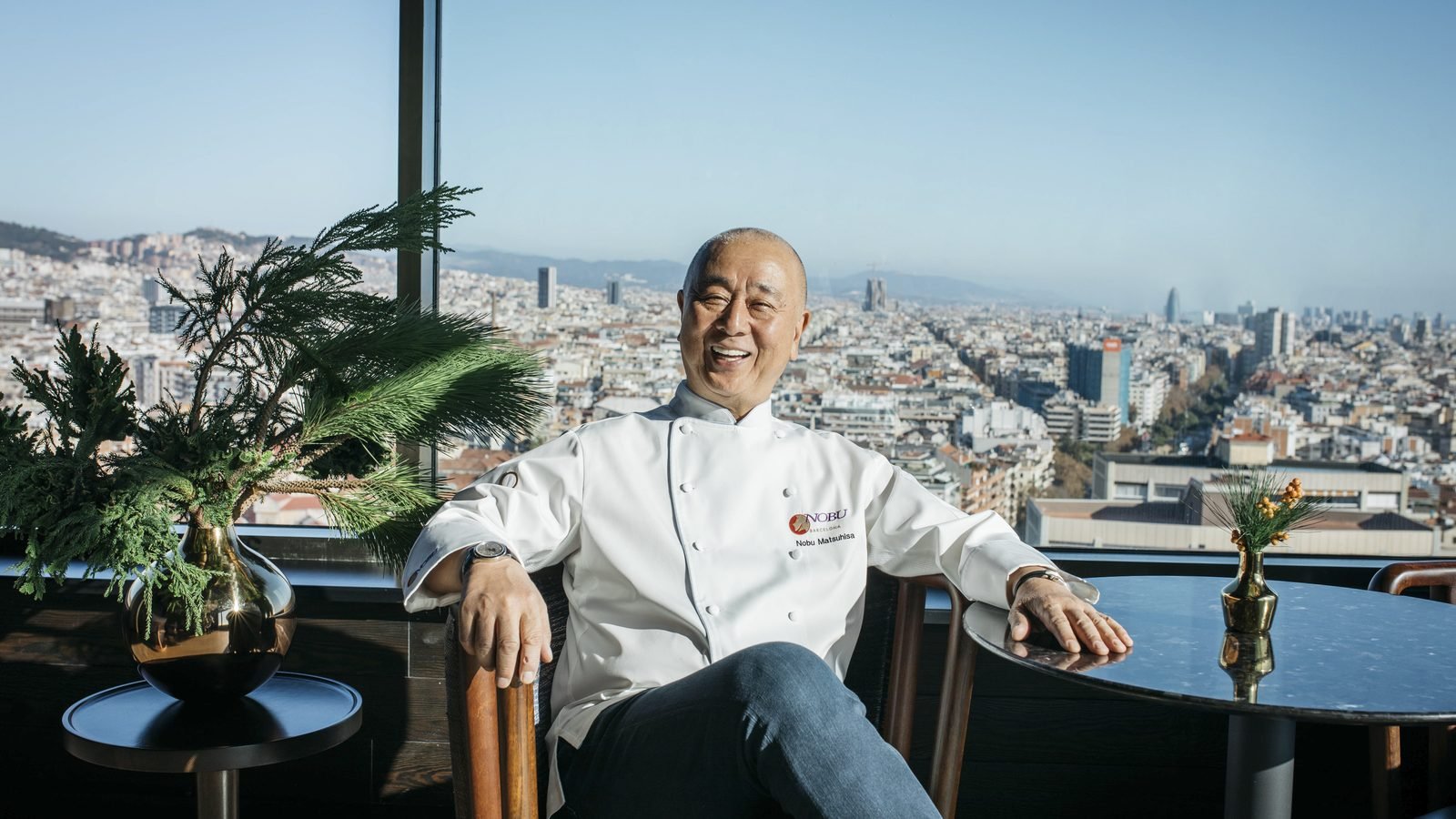 El chef Nobu Matsuhisa sonríe sentado junto a una ventana con Barcelona al fondo