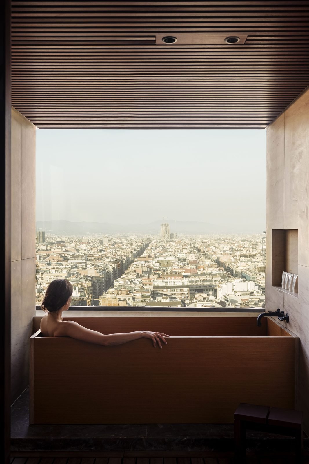 Una mujer en una tina de madera frente a una ventana con vista a la ciudad