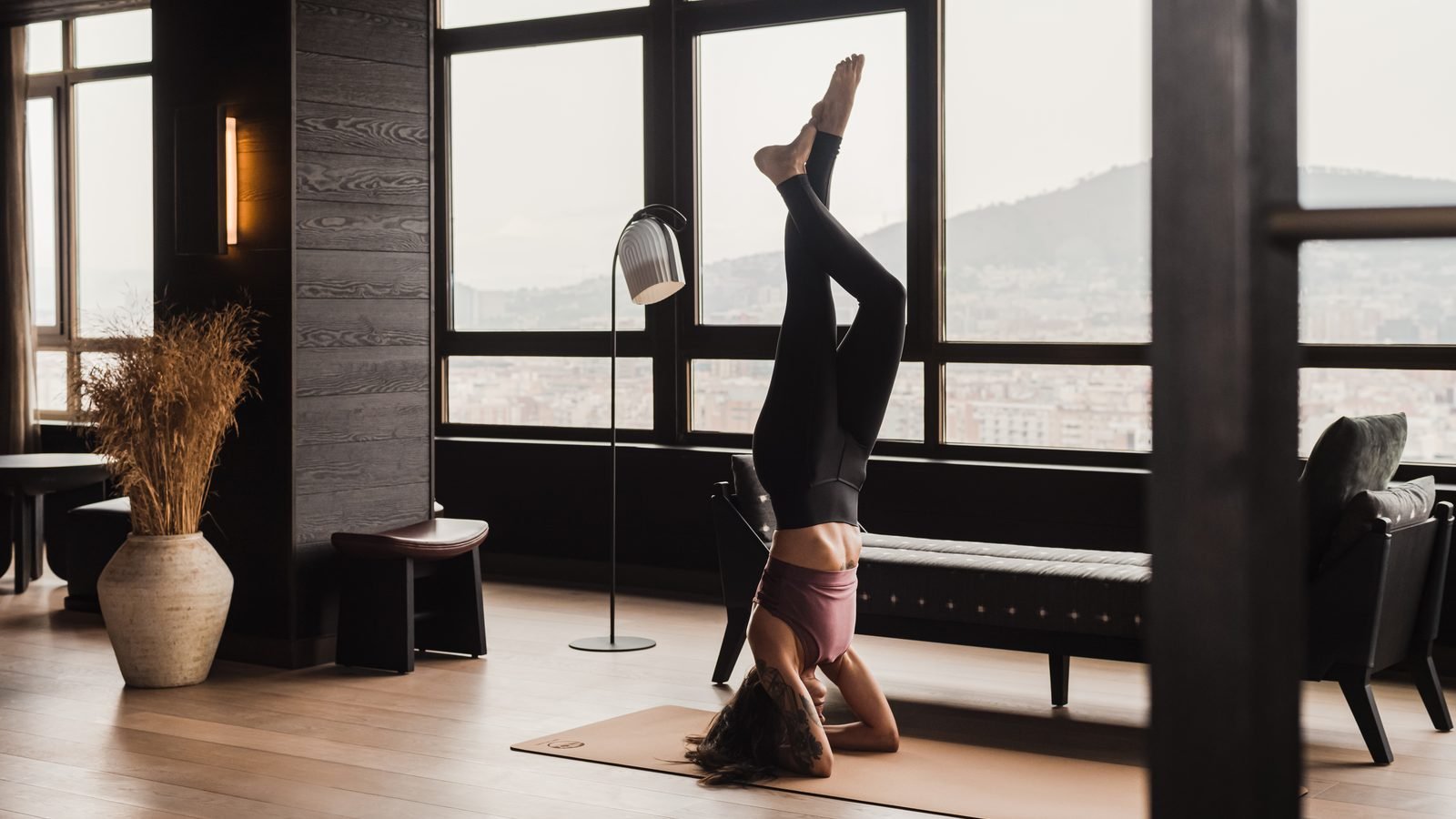 Una mujer hace una postura de yoga sobre la cabeza frente a un ventanal con la ciudad