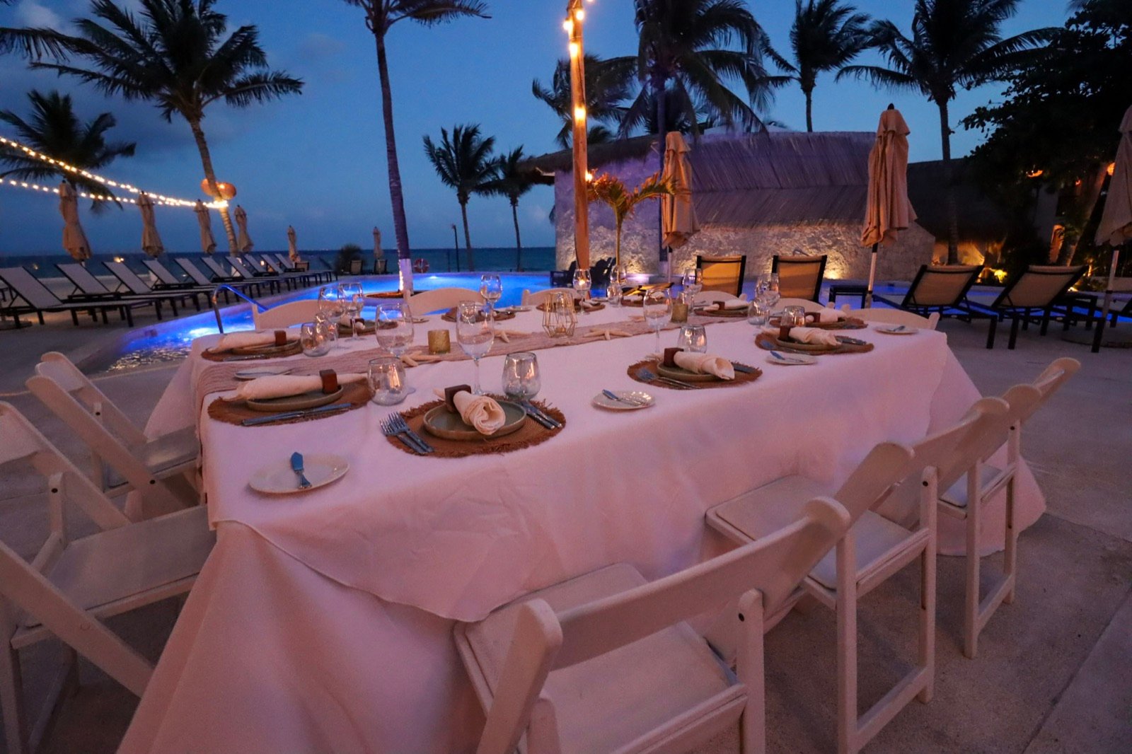 Mesa larga de cena junto a la alberca iluminada, de noche, con palmeras y foquitos