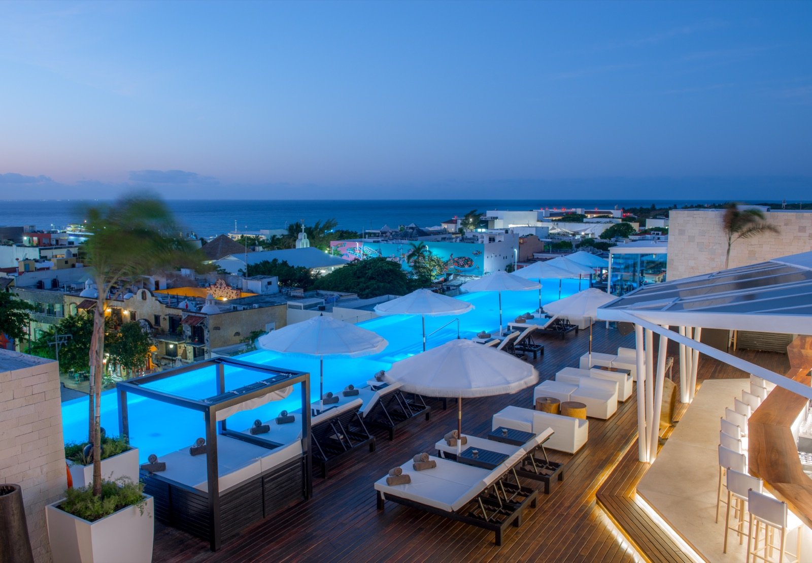 El rooftop de noche, con la alberca iluminada de azul y Playa del Carmen alrededor